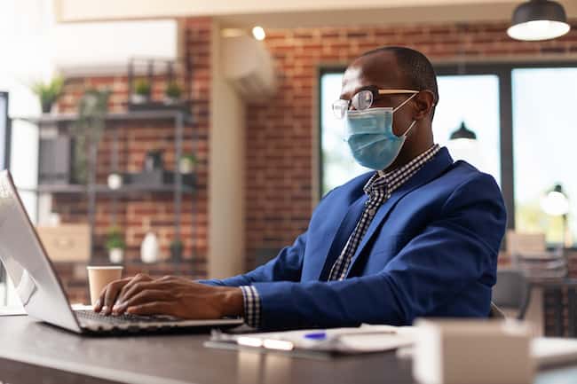 Человек смотрит на экран ноутбука, чтобы спланировать бизнес-проект во время пандемии коронавируса. Предприниматель в маске использует компьютер для работы над маркетинговой стратегией в стартап-офисе.